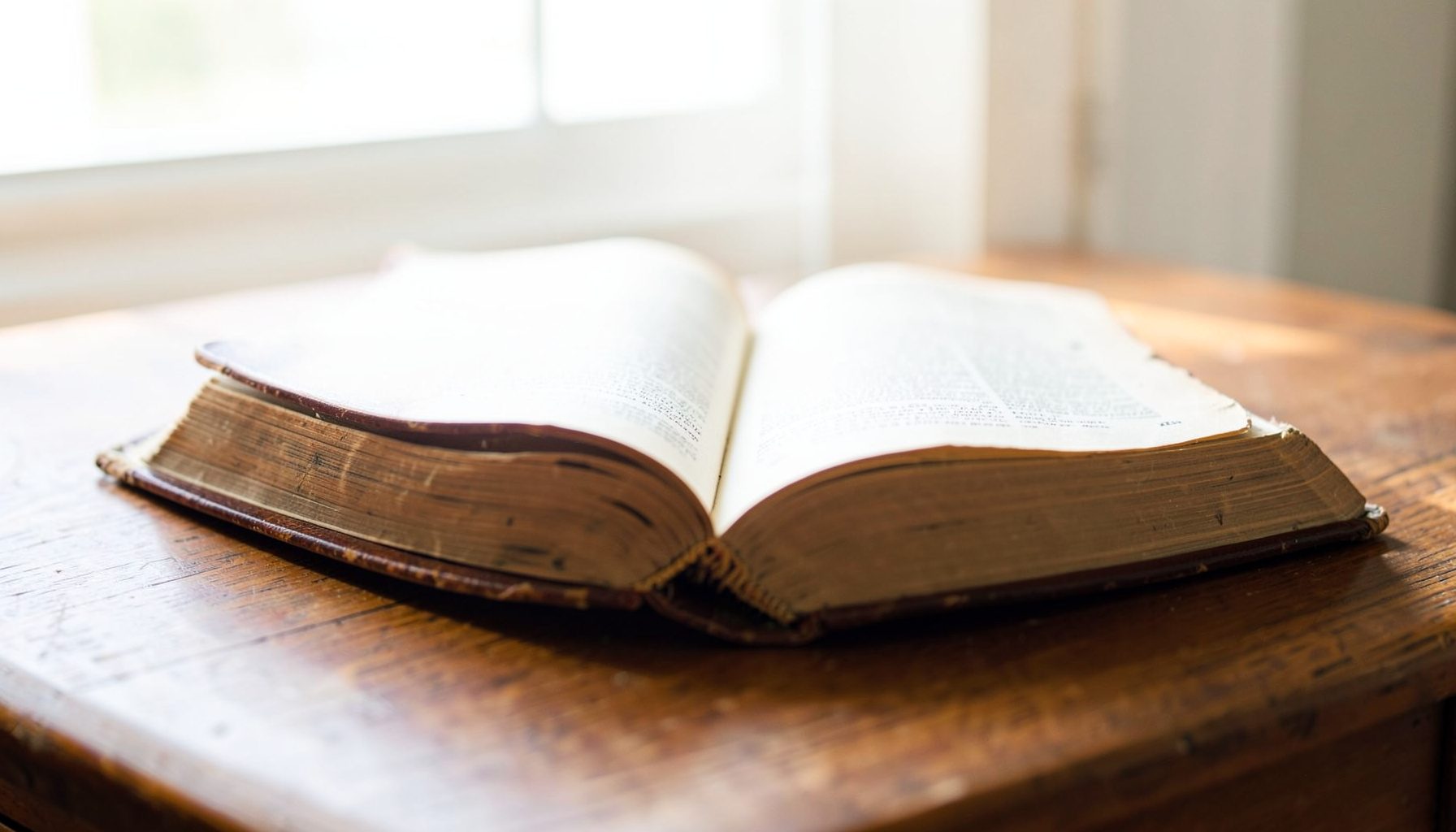 An open antique Bible on a wooden desk, warm brown leather cover with gold-edged pages, soft natural window light