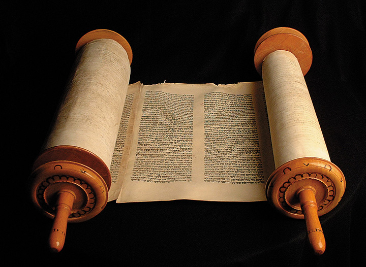 An open scroll with wooden handles resting on a dark background, its handwritten script columns rendered in warm beige and brown tones, evoking careful historical study